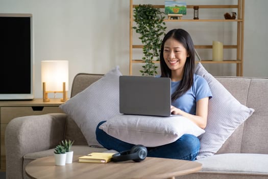 Happy cheerful young asian woman sitting on sofa with laptop, smart beautiful young asian woman chilling in house with