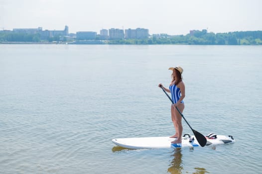A red-haired woman in a striped swimsuit and a straw hat floats on a paddle board on the lake
