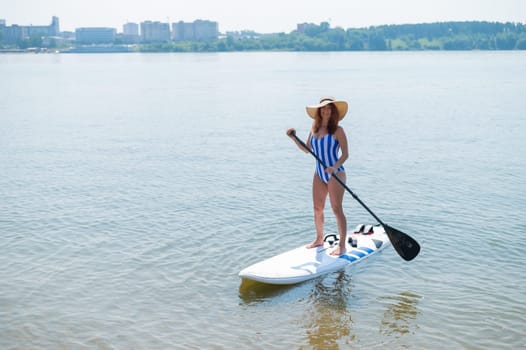 A red-haired woman in a striped swimsuit and a straw hat floats on a paddle board on the lake