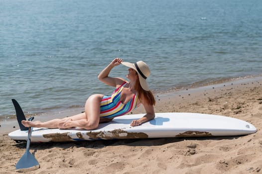A woman in a swimsuit and a hat is posing lying on a sup board on the beach.