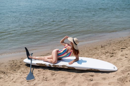 A woman in a swimsuit and a hat is posing lying on a sup board on the beach.