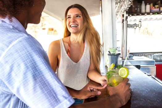 Young blond woman having cocktails and talking with black male friend at beach bar.