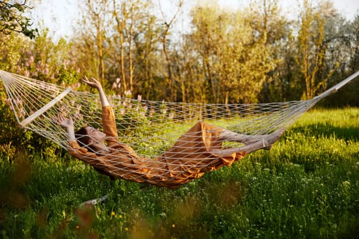 a funny woman is resting in nature lying in a mesh hammock in a long orange dress, raising her hands up, enjoying the rays of the setting sun on a warm summer day