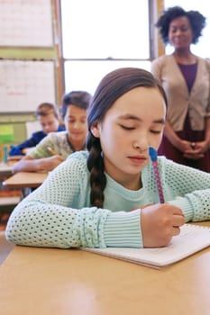 Shes focused on this test. a young girl sitting in class with her teacher and classmates blurred in the background.