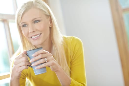 A beautiful woman smiling and sitting near a window inside a house. Happy attractive blonde woman chilling drinking coffee. Happy female relaxing and daydreaming at home off the grid in a sunroom