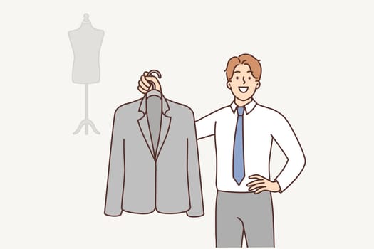 Man fashion designer is holding business suit on hanger providing tailoring services. Guy working as fashion designer demonstrates new or repaired jacket and looks at camera smiling