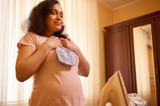 Delightful pregnant woman with big belly holds an ironed bodysuit for her future newborn baby.