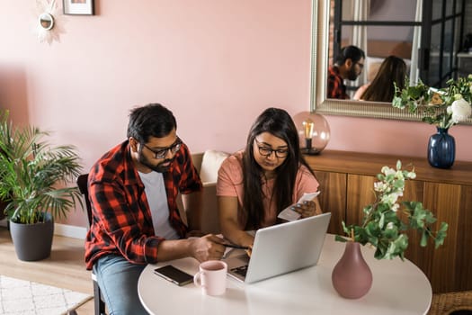 Young millennial indian husband and wife doing domestic paperwork, accounting job and reviewing paper bills, receipts at laptop computer, using online calculator and paying mortgage rent fees on internet - economic crisis concept