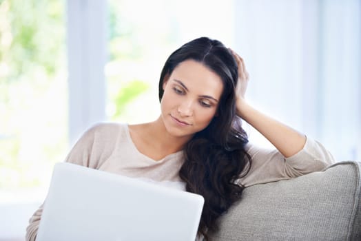 Surfing the web at home. A young woman sitting on her sofa and looking at her laptop screen while relaxing.