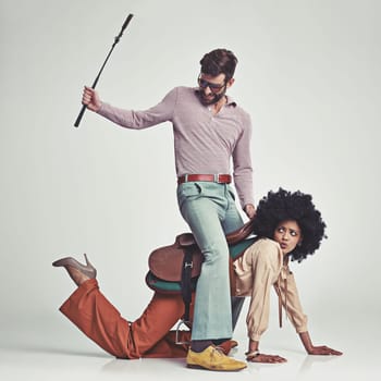 Hes in charge now. A studio shot of an attractive man in 70s wear riding a young woman wearing a saddle while using a riding crop.