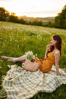 happy redhead woman sits in a field of daisies on a plaid during sunset and enjoys nature