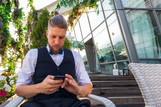 Young hipster in a white shirt, black vest looks into a smartphone while sitting in a penthouse