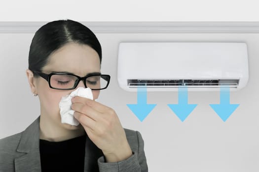 A woman with a tissue to her nose is having a cold and feeling unwell due to the air conditioner in the office