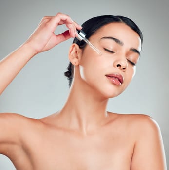 A beautiful mixed race woman applying a soothing face serum to her radiant smooth face while posing against a blue background. Hispanic woman with flawless skin using essential oil