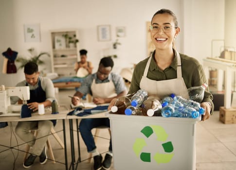 Cheerful seamstress holding a bucket of recycled plastic bottles. Mixed race designer holding bin of renewable plastic bottles. Young tailor recycling plastic bottles from her studio
