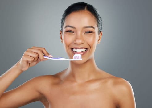Oral hygiene is a big thing. a beautiful young woman brushing her teeth while standing against a grey background.