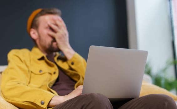 Man sitting on sofa with laptop, showing facepalm gesture with hand on face. Focus is on laptop, which suggests that the man may be experiencing frustration or disappointment related to the technology
