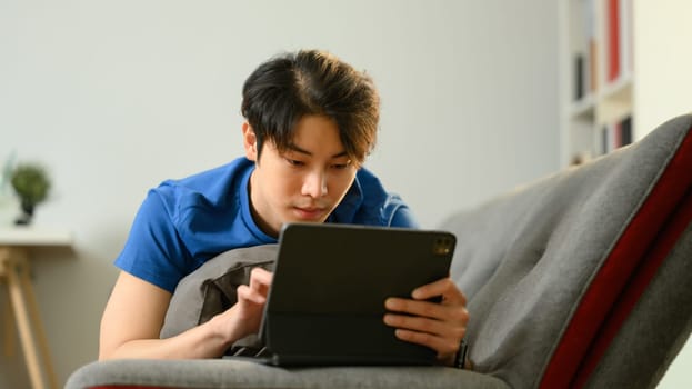 Casual asian man lying on couch hand using digital tablet. People, technology and lifestyle.
