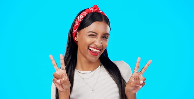 Face, funny and Indian woman with peace sign, confident and happiness on a blue studio background. Portrait, female and lady with smile, silly and goofy with wink, joyful and cheerful with excitement