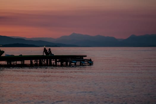 Sunset time on river. Orange sunset landscape with pier and men. Vivid orange sky view. Romantic evening sunset in tropics. Rustic wooden pier and boat silhouette. Sunset watchers. Sun over mountain