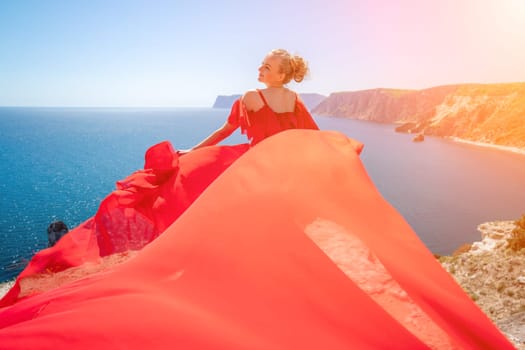 Blonde with long hair on a sunny seashore in a red flowing dress, back view, silk fabric waving in the wind. Against the backdrop of the blue sky and mountains on the seashore.
