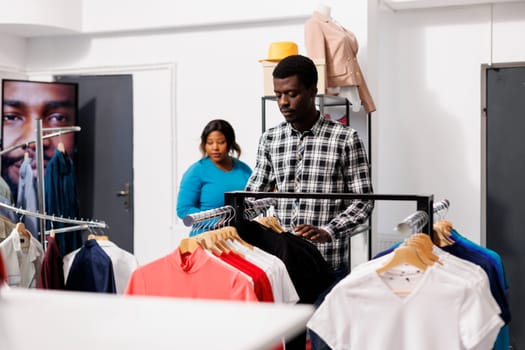 African american man looking at new fashion collection in modern boutique, shopping for trendy merchandise. Stylish customer checking clothes material and price in clothing store. Fashion concept