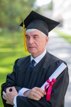 Portrait of an elderly man in a graduation gown and with a diploma in his hands outdoors. Vertical.