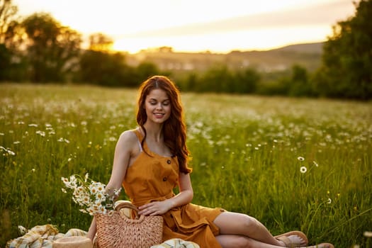 a happy woman in an orange dress sits on a plaid in a chamomile field at sunset and laughs