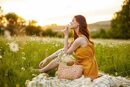 beautiful, happy woman in an orange dress is resting sitting on a plaid in a chamomile field