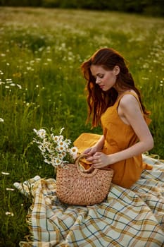 happy redhead woman sits in a field of daisies on a plaid during sunset and enjoys nature