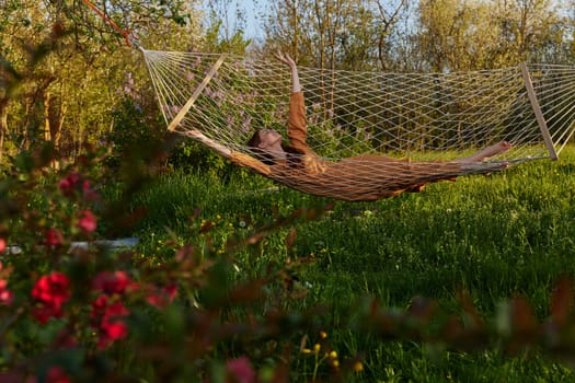 a happy woman in a long orange dress is relaxing in nature lying in a mesh hammock enjoying summer and vacation in the country surrounded by green foliage, happily lifting her legs
