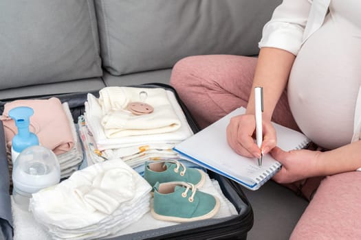 Pregnant woman writing packing list for maternity hospital sitting on a couch next to suitcase