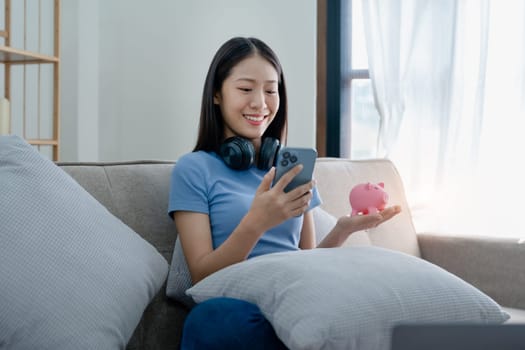 Asian young woman holding a piggy bank and using smartphone mobile, Savings concepts.