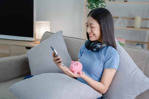 Asian young woman holding a piggy bank and using smartphone mobile, Savings concepts.