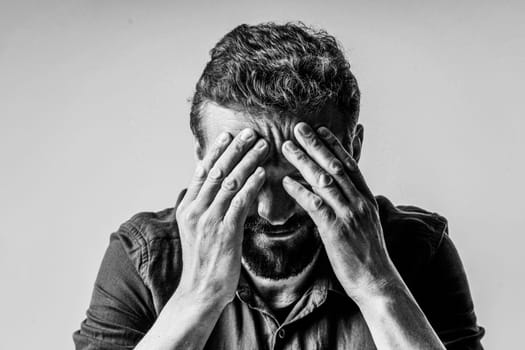 Black and white photo of a man looking extremely sad and depressed. He sitting on white background and has face covered with both hands, showing the despair and hopelessness he feels.