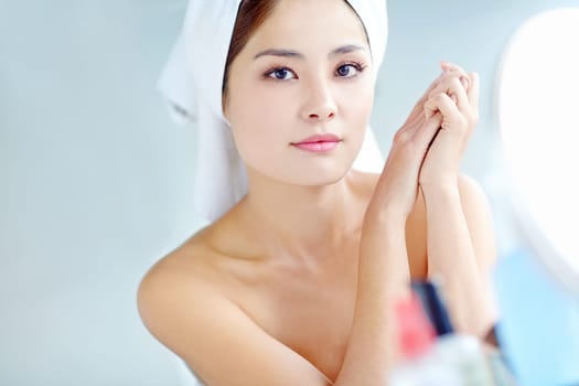 Moisturizing her hands. A beautiful young Asian woman smiling while sitting in front of a mirror with a towel on her head.