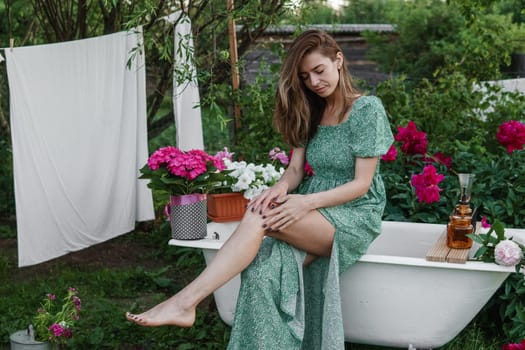 A woman is sitting on a cast-iron bathtub in the courtyard of a country house next to a bush of flowering peonies. The concept of summer, country life.