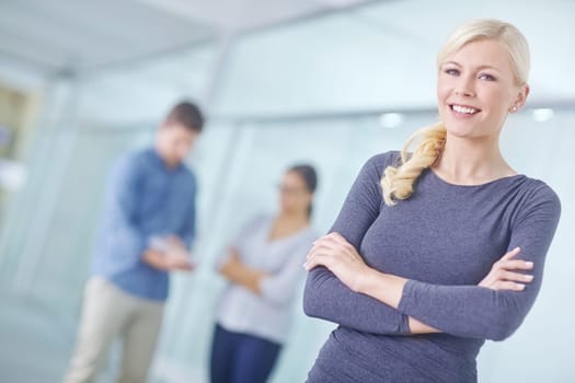 Portrait of a smiling businesswoman standing with her arms crossed and looking at the camera while her colleagues work in the background. One female entrepreneur leader ready for business success