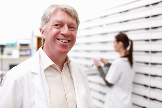 Smiling pharmacist with colleague working. Portrait of mature pharmacist smiling with female colleague working in background.