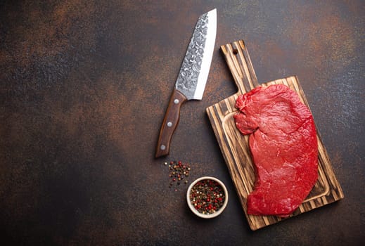 Raw uncooked top round beef steak on wooden cutting board with big kitchen knife and pepper on dark brown rustic stone background top view, cooking meat steak with copy space