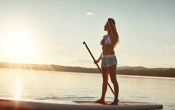 All I need is my board and sun. an attractive young woman paddle boarding on a lake.
