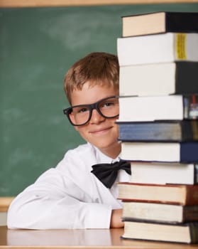 He has a thirst for knowledge. A young boy wearing glasses and a bow-tie smiling at the camera from behind a stack of books.