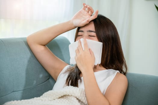 Sick woman sitting under blanket on sofa and sneeze with tissue paper in living room