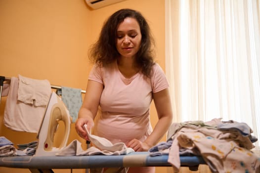 Expectant mother, pregnant woman ironing and folding newborn clothes, enjoying future maternity and carefree pregnancy