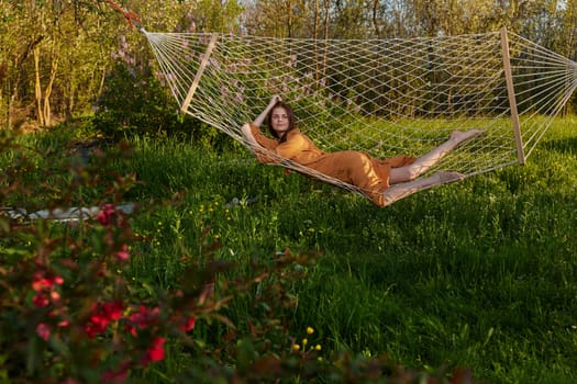 a happy woman in a long orange dress is relaxing in nature lying in a mesh hammock enjoying summer and vacation in the country surrounded by green foliage