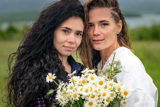 Two women enjoy nature in a field of daisies. Girlfriends hugging hold a bouquet of daisies and look at the camera.