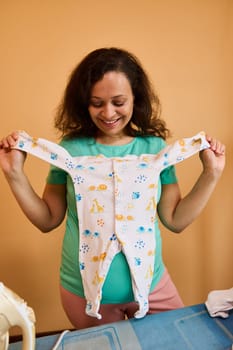 Smiling pregnant woman holding jumpsuit for newborn baby, standing by ironing desk, preparing bag for maternity hospital