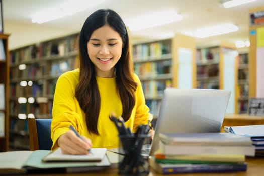 Beautiful Asian college woman preparing report, doing assignments on laptop in a library