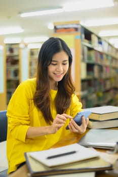Portrait of smiling young female student sitting at table in library with textbooks and using smartphone
