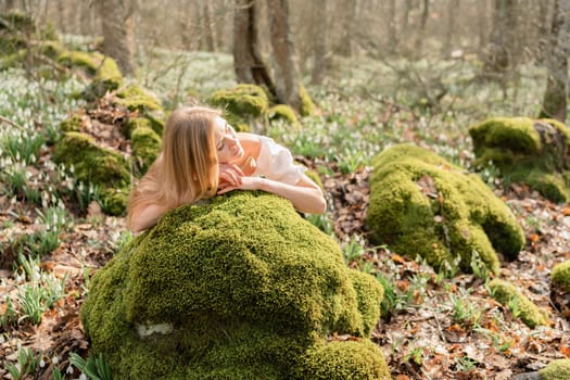 Snowdrops galanthus blond. A girl in a white dress lay down on a stone in the moss in a meadow with snowdrops in a spring forest